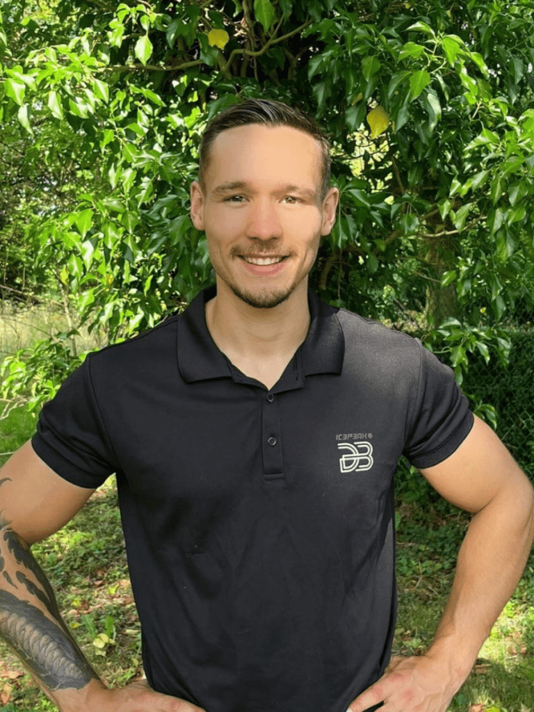 Un homme vêtu d'un polo noir se tient dehors, devant un feuillage vert, souriant, les mains sur les hanches, respirant l'assurance typique d'un chef d'équipe pédagogique. fitness academy luxembourg par david blondiau