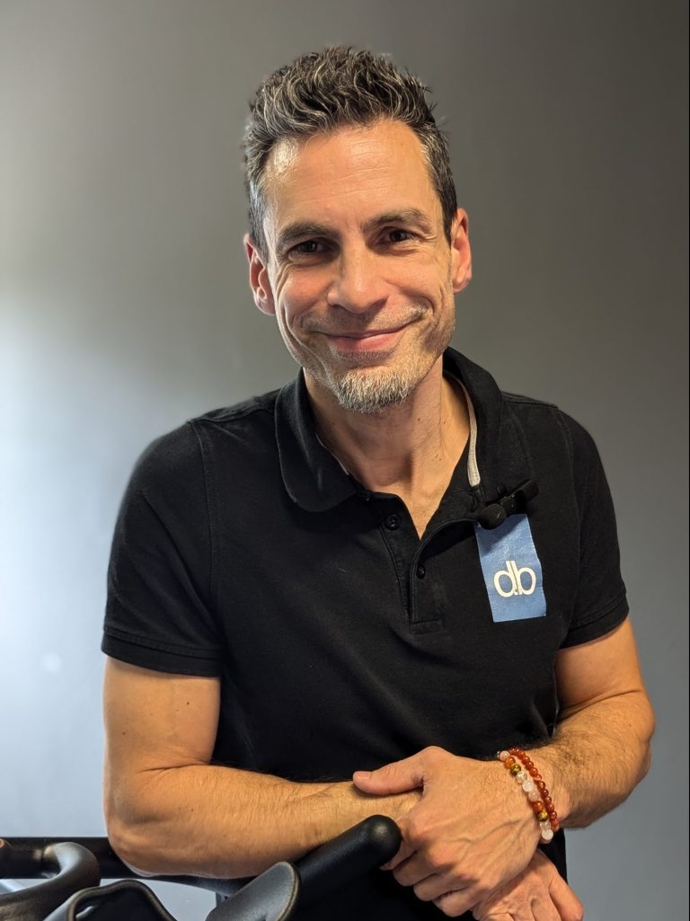 Un homme aux cheveux poivre et sel et portant une barbiche porte un polo noir avec le logo « db ». Il s'appuie sur un équipement de gym, sourit à la caméra, comme s'il venait de terminer l'entraînement de son équipe. Fitness Academy by David Blondiau